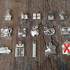 May include: A collection of silver-toned charms with school-themed designs. Charms include a school bus, apple, books, pencils, a calculator, and a ruler. Some charms have text like "Best Teacher Ever" and "#1 Teacher".