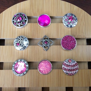 May include: Nine pink and silver snap buttons with various designs, including a sun, a flower, and a star. The buttons are arranged in three rows on a wooden surface.