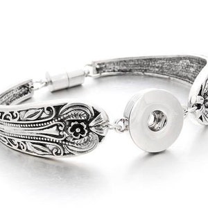 May include: A silver bracelet with an ornate design and a snap closure. The bracelet is made from a repurposed spoon.