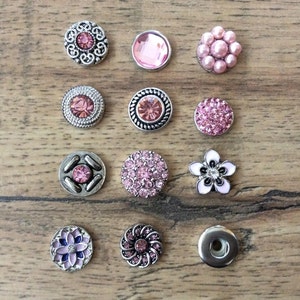 May include: Twelve assorted snap buttons with pink and white accents. The buttons feature various designs, including flowers, swirls, and geometric shapes.