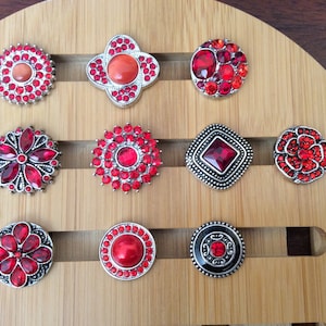 May include: Twelve red rhinestone snap buttons in various shapes and designs, including a flower, a circle, a square, and a clover. The buttons are displayed on a wooden surface.
