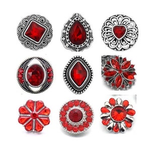 May include: Nine red rhinestone snap buttons with silver metal frames. The buttons feature various designs, including a square, teardrop, heart, oval, flower, and circle.