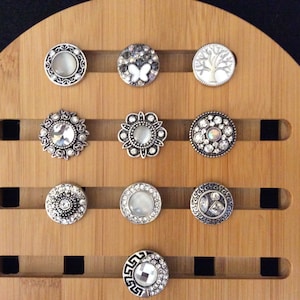 May include: A wooden display stand with 10 round, silver-toned snap buttons with various designs, including a butterfly, a tree, and geometric patterns.