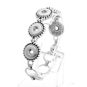 May include: Silver-toned bracelet featuring three large, round, snap-on style charms with a decorative edge. The bracelet is connected by smaller, circular links and is displayed on a clear stand. The design is intricate and detailed.