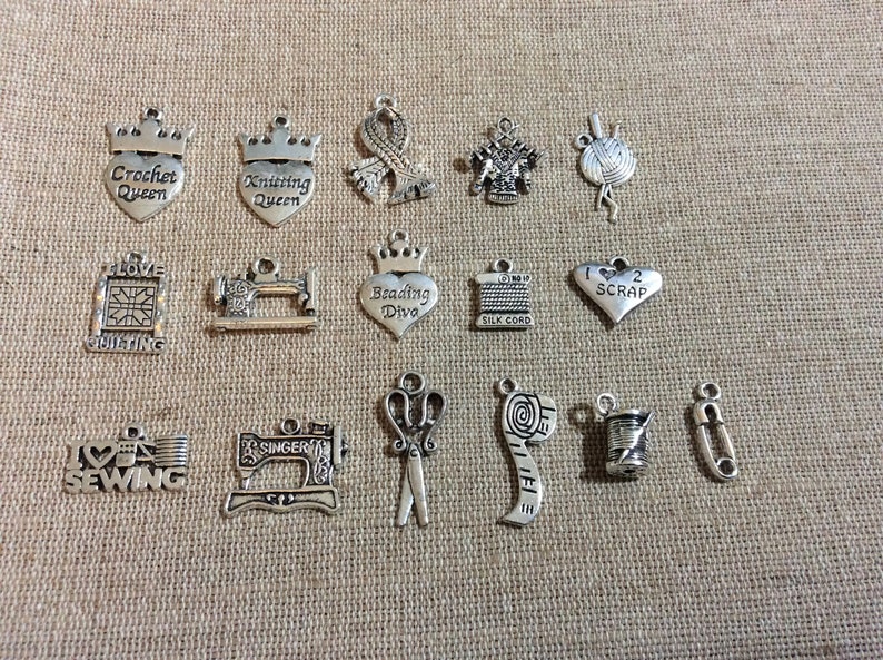 Puede incluir: Una colecci&oacute;n de 15 colgantes de amuleto de plata con varios temas de artesan&iacute;a, que incluyen costura, tejido, ganchillo, acolchado y abalorios. Los amuletos son todos en forma de corazones, coronas u otras herramientas de artesan&iacute;a.