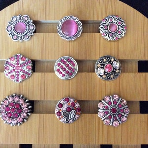May include: Assortment of 10 pink and silver snap jewelry buttons. The buttons feature various designs including a flower, a butterfly, and a swirl pattern.