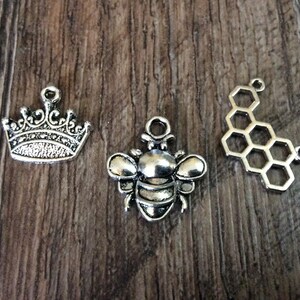 May include: Three silver-colored charms on a wooden surface. The charms include a detailed crown, a bee, and a honeycomb shape. These charms are likely for jewelry making or other craft projects.