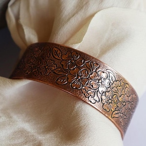 May include: A copper bracelet featuring an embossed floral design. The bracelet is a wide band with a raised pattern of flowers and leaves.