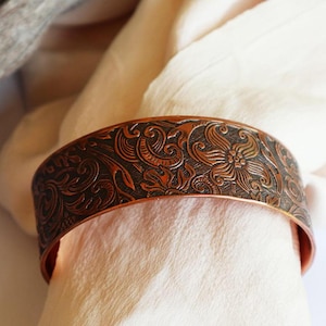 May include: A copper bracelet featuring an elaborate floral pattern. The bracelet is wide and has a smooth, polished finish.