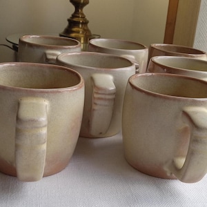 May include: A set of six beige ceramic mugs with a light brown glaze. The mugs have a rounded shape and a handle on each side.