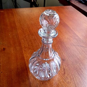 May include: A clear glass decanter with a round, patterned stopper. The decanter has a bulbous base and a slender neck. The stopper is spherical with a detailed, etched design. The glass is clear, reflecting light.