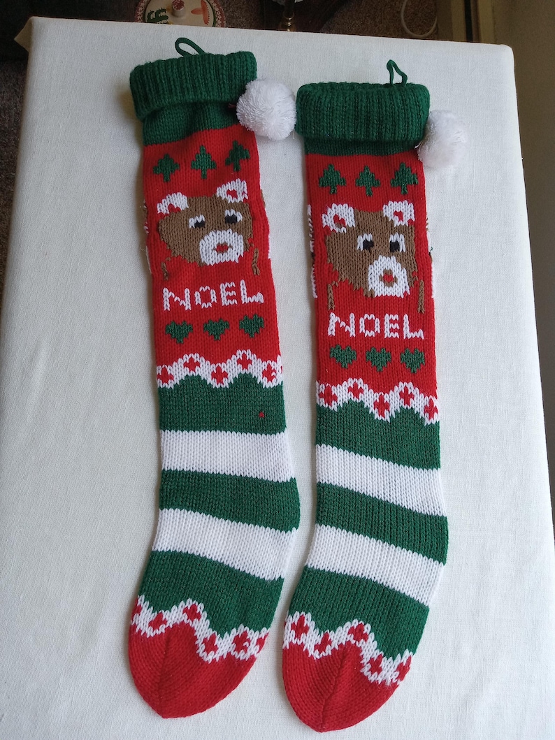 May include: Two red and green Christmas stockings with a reindeer design and the word "NOEL". The stockings have white and green stripes at the bottom and a white pom-pom at the top. A festive holiday decoration.