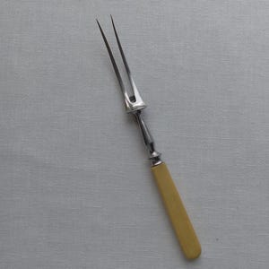 May include: A vintage carving fork with a two-pronged stainless steel head and a cream-colored handle. The handle is made of a smooth, solid material. The fork is designed for serving meats.