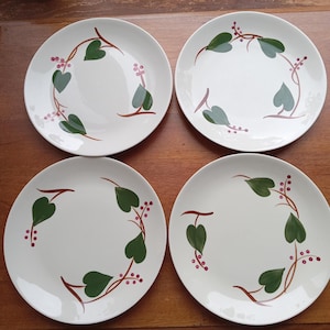 May include: Four white ceramic plates with a green leaf and berry design. The leaves are heart-shaped and the berries are small and red. The plates are arranged on a wooden surface.
