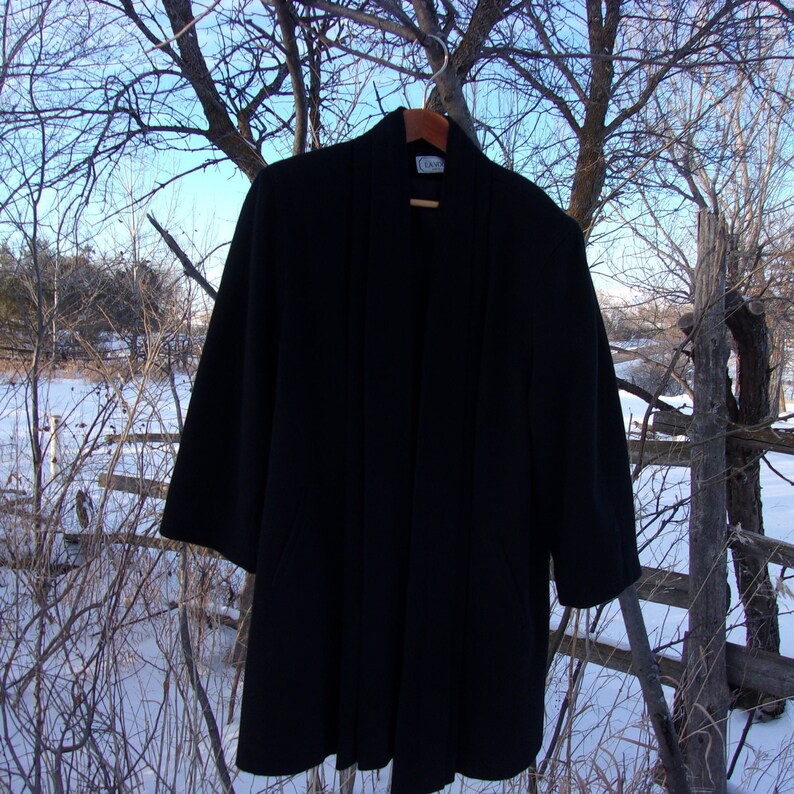 May include: A black long-sleeved cardigan with a shawl collar and a simple design. The cardigan is hanging on a wooden hanger against a snowy background.