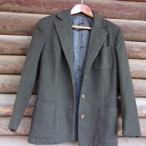 May include: A dark green blazer with a two-button closure and two front pockets. The blazer is hanging on a wooden hanger against a wooden background. The blazer has a notched lapel and long sleeves.
