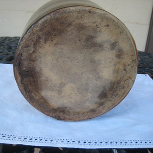 May include: The bottom of a large, brown stoneware crock with a glazed finish. The crock has a wide, round base and a slightly flared lip.