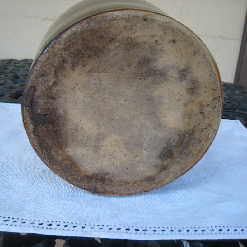 May include: The bottom of a large, brown stoneware crock with a glazed finish. The crock has a wide, round base and a slightly flared lip.