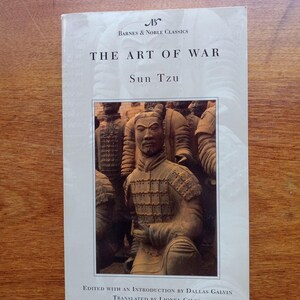 May include: A paperback book titled "THE ART OF WAR" by Sun Tzu, published by Barnes & Noble Classics. The cover features a photograph of a terracotta warrior statue. The book is edited by Dallas Galvin and translated by Lionel Giles.