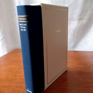 May include: A hardcover book titled "LINCOLN Speeches and Writings 1832-1858" with a dark blue spine and gold lettering. The adjacent book has a cream-colored cover with a gold emblem. The books are on a wooden surface.