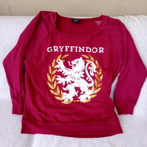 May include: A maroon sweatshirt with long sleeves. The front features the word "GRYFFINDOR" in white block letters above a white lion emblem encircled by a gold laurel wreath. The sweatshirt is made of a soft material.