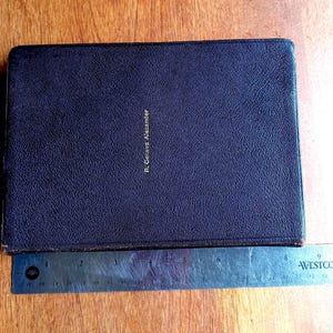 May include: A rectangular dark brown book with a textured cover and the name R. Geneva Alexander embossed in gold lettering. A metal ruler is positioned along the bottom edge, showing the book has a width of approximately 9 inches.