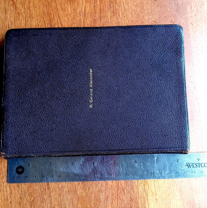 May include: A rectangular dark brown book with a textured cover and the name R. Geneva Alexander embossed in gold lettering. A metal ruler is positioned along the bottom edge, showing the book has a width of approximately 9 inches.