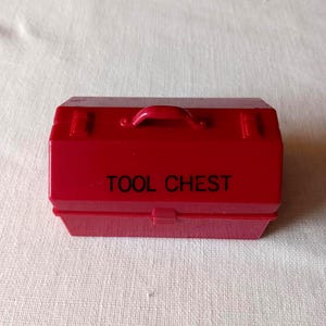 May include: A small, red tool chest with a handle and the words "TOOL CHEST" printed in black. The chest has a rectangular shape and a hinged lid, suggesting it is designed for storing tools.