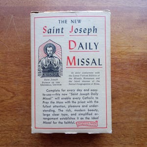 May include: The cover of a vintage book titled "The New Saint Joseph Daily Missal". The cover features a drawing of Saint Joseph in a red, white, and blue robe, with a halo and a crown. The text on the cover is in black and red.