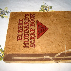 May include: A square scrapbook with a tan, textured cover featuring the text ELBERT HUBBARD'S SCRAP BOOK in dark brown lettering and a red triangular graphic. The book is bound with a thin, light-colored fabric tie knotted on the front cover.