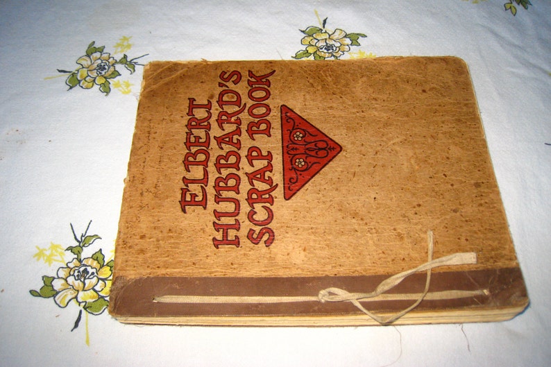 May include: A square scrapbook with a tan, textured cover featuring the text ELBERT HUBBARD'S SCRAP BOOK in dark brown lettering and a red triangular graphic. The book is bound with a thin, light-colored fabric tie knotted on the front cover.