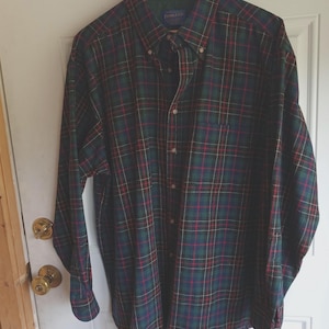May include: A long-sleeve button-down shirt featuring a dark green, navy blue, and red plaid pattern. The shirt has a pointed collar, a single chest pocket on the left side, and a row of buttons down the front. The Pendleton brand label is visible.