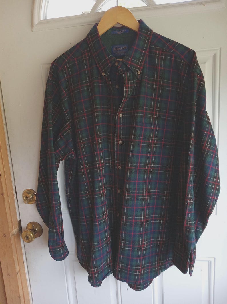 May include: A long-sleeve button-down shirt featuring a dark green, navy blue, and red plaid pattern. The shirt has a pointed collar, a single chest pocket on the left side, and a row of buttons down the front. The Pendleton brand label is visible.