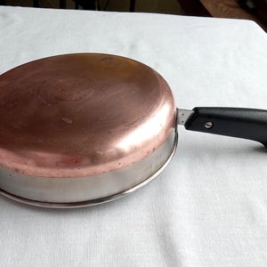 May include: A copper-bottomed frying pan with a stainless steel rim and a black handle. The pan is round and has a shiny copper surface on the bottom. The handle is black and has a loop for hanging. Kitchenware.