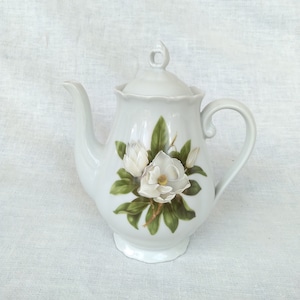 May include: White porcelain teapot with a floral design featuring white magnolia blossoms and green leaves. The teapot has a lid and a handle.