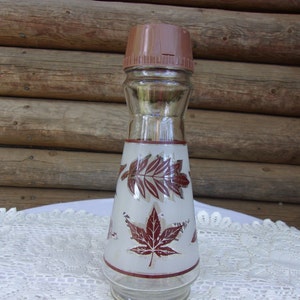 May include: A clear glass spice grinder with a brown plastic lid. The glass body features a frosted design with red and gold leaf patterns. A red band accents the base and the top of the frosted section. The grinder is approximately 7 inches tall.