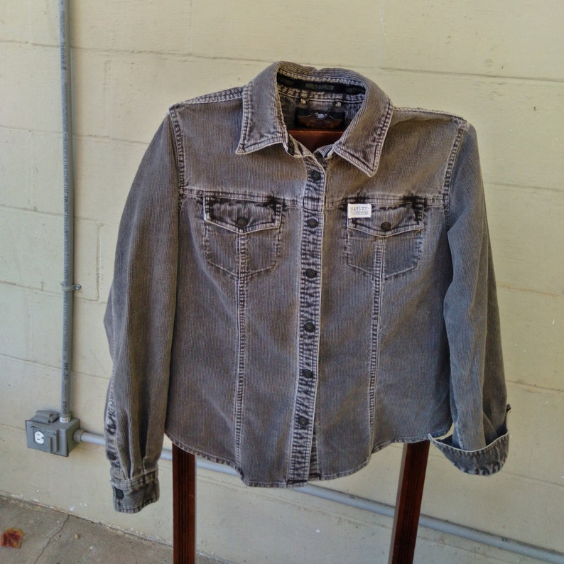 May include: A gray corduroy button-up shirt with a long sleeve and two chest pockets. The shirt has a small white label with the text "Barneys New York" on the front.
