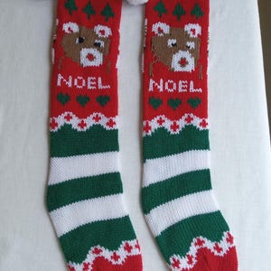 May include: Pair of Christmas stockings with a red, green, and white color scheme. Each stocking features a reindeer design, the word "NOEL", and a striped pattern. The top has a green cuff with a white pom-pom.
