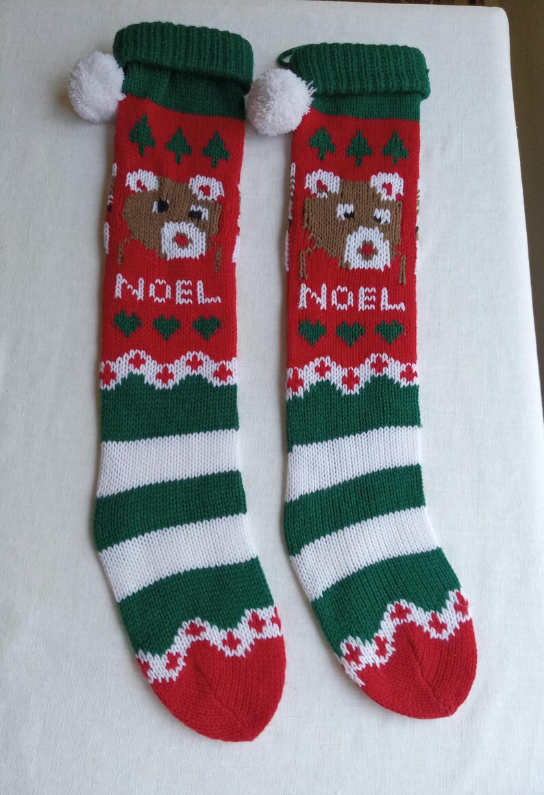 May include: Pair of Christmas stockings with a red, green, and white color scheme. Each stocking features a reindeer design, the word "NOEL", and a striped pattern. The top has a green cuff with a white pom-pom.