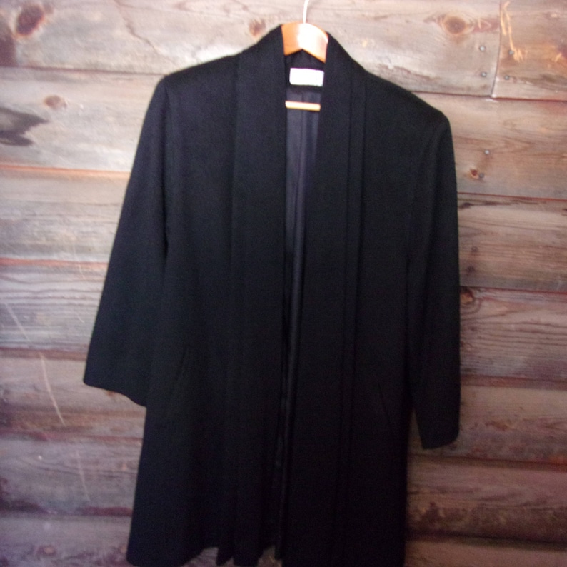 May include: A black long-sleeved jacket with a shawl collar and a long, flowing design. The jacket is hanging on a wooden hanger against a wood panel background.