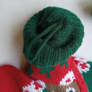 May include: Close-up of a green knitted hat with a drawstring. The hat is part of a Christmas stocking with red, white, and green knitted details, including a reindeer face. The stocking is made of soft yarn.