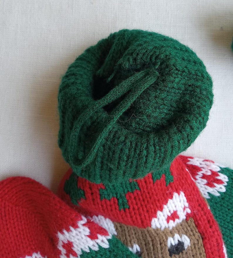 May include: Close-up of a green knitted hat with a drawstring. The hat is part of a Christmas stocking with red, white, and green knitted details, including a reindeer face. The stocking is made of soft yarn.