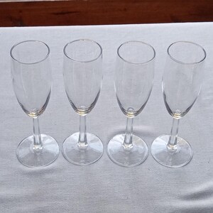 May include: Four clear champagne flutes with slender stems and round bases are arranged in a row. The glasses are empty and sit on a white surface, with a wooden background visible.