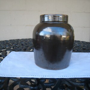 May include: A dark brown glazed stoneware crock with a wide mouth and a rounded body. The crock is sitting on a white doily.
