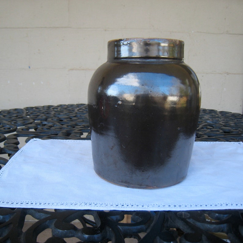 May include: A dark brown glazed stoneware crock with a wide mouth and a rounded body. The crock is sitting on a white doily.
