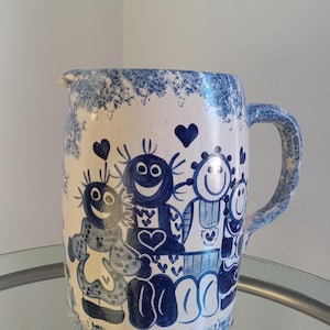 May include: A white ceramic pitcher with a blue and white design featuring four smiling figures. The figures are wearing blue and white clothing and have hearts on their chests. The pitcher has a handle and a speckled blue glaze.