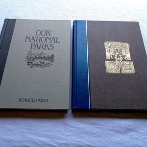 May include: Two hardcover books placed side by side. The left book is gray with a black spine, featuring the text "OUR NATIONAL PARKS" and "READERS DIGEST" with a landscape illustration. The right book is brown with a blue spine and a gold illustration.