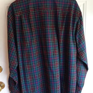 May include: A long-sleeve button-down shirt featuring a plaid pattern in dark green, navy blue, and red with thin white accent lines. The shirt has a standard collar, a back yoke, and is displayed hanging on a wooden hanger against a white door.