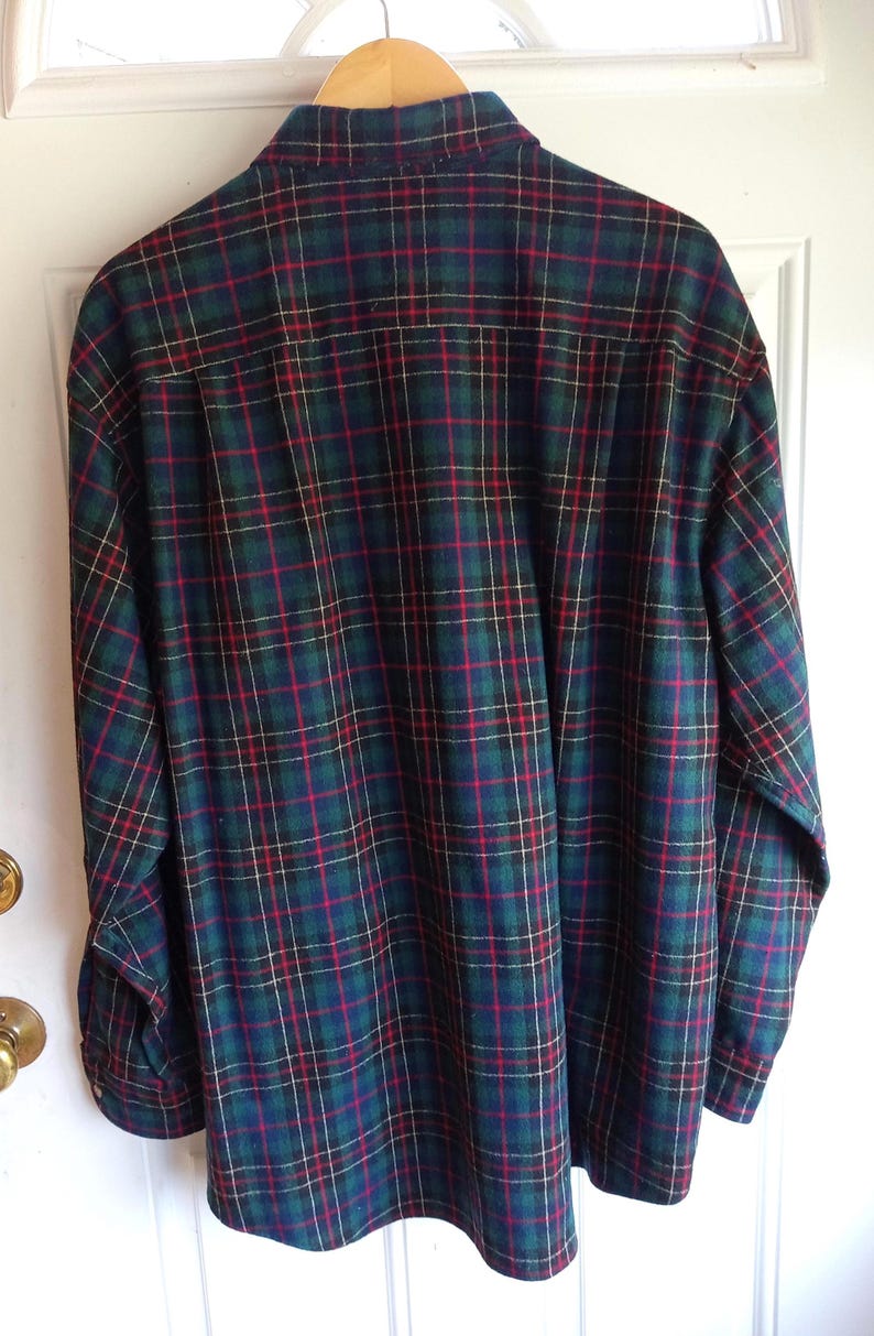 May include: A long-sleeve button-down shirt featuring a plaid pattern in dark green, navy blue, and red with thin white accent lines. The shirt has a standard collar, a back yoke, and is displayed hanging on a wooden hanger against a white door.