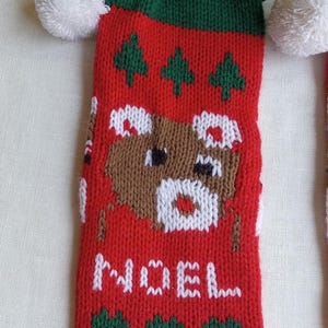 May include: Red Christmas stocking with a green cuff and white pom-pom. The stocking features a knitted design with a brown bear, green trees, and the word "NOEL" in white. A festive holiday decoration.